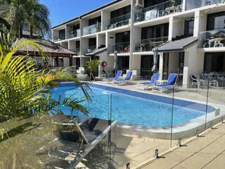 Outdoor swimming pool area with blue lounge chairs, surrounded by a glass fence, in front of a multi-story apartment building.