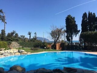Outdoor swimming pool surrounded by trees and shrubs, with distant mountains and a clear blue sky in the background.