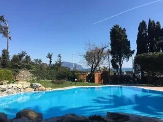 Outdoor swimming pool surrounded by trees and shrubs, with distant mountains and a clear blue sky in the background.