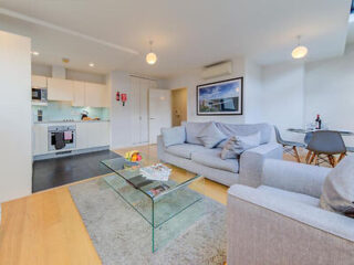 Modern living room with grey sofas, glass coffee table, and a kitchen in the background. A round dining table with chairs is on the right. Bright lighting and minimalist decor.