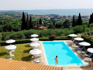 A person stands in a rectangular outdoor pool surrounded by sun loungers and umbrellas. The landscape includes trees and a distant view of a body of water and town.