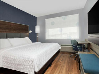 Hotel room with a large bed, dark wooden headboard, desk with chair, and a wall-mounted TV. The window has a sheer white blind, and the floor is wood.