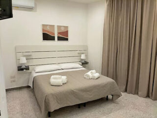 A neat bedroom with a double bed, beige bedspread, and rolled towels on top. The wall has two abstract paintings above the headboard, and long curtains hang on the right side.