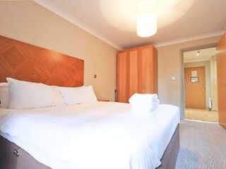 A neatly made bed with white linens in a hotel room; a wooden wardrobe and a closed door are in the background.