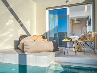 A beige outdoor lounge area with a bean bag and wicker chair next to a small pool. A table holds fruits and drinks. Glass doors open to a modern interior space.