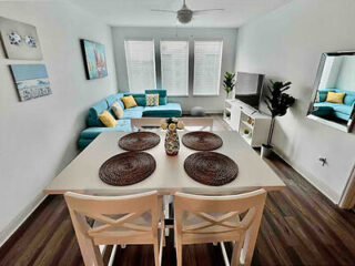 Modern living room with a teal corner sofa, wall art, TV, and dining table set for four with wicker placemats. Three large windows bring in natural light.