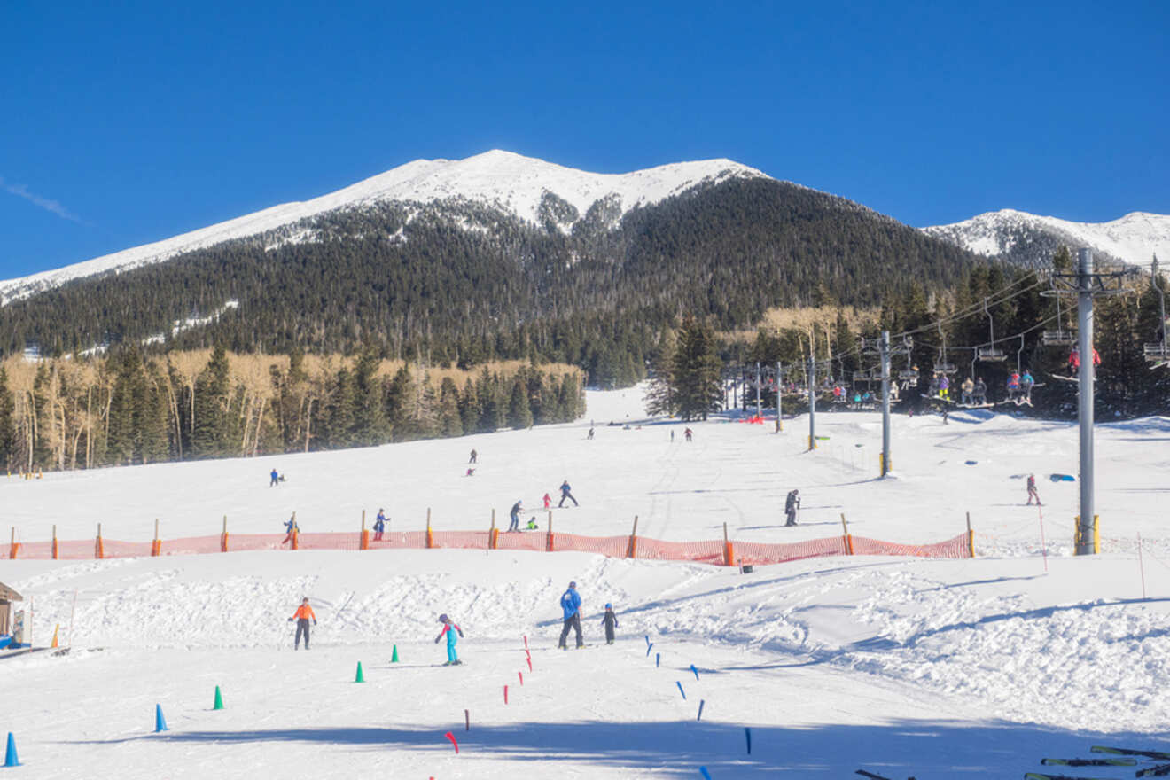 Skiers on a snow-covered slope with a chairlift and mountain in the background under a clear blue sky.