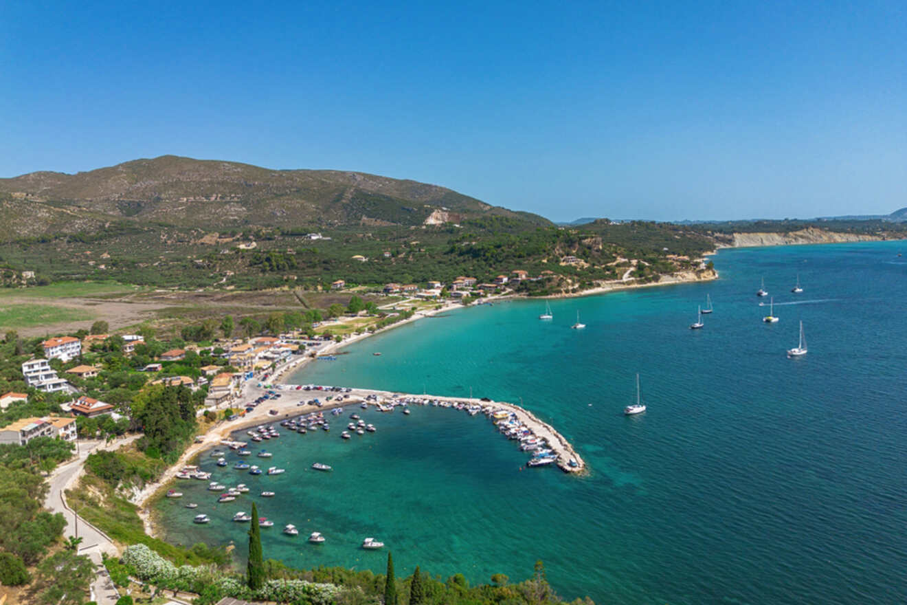 Aerial view of a coastal village with a small harbor, boats anchored in turquoise waters, surrounded by hills and sparse buildings under a clear blue sky.