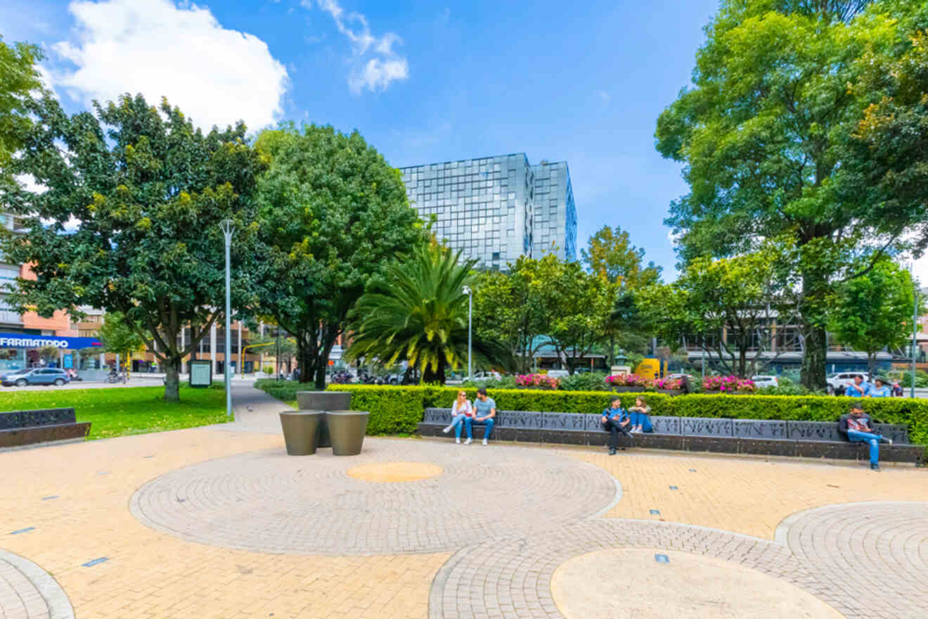 Urban park scene with people sitting on benches, surrounded by trees and plants. Modern buildings are visible in the background.