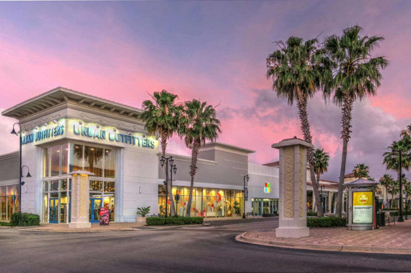 Shopping center with an Urban Outfitters store, palm trees, and a colorful sky at sunset.