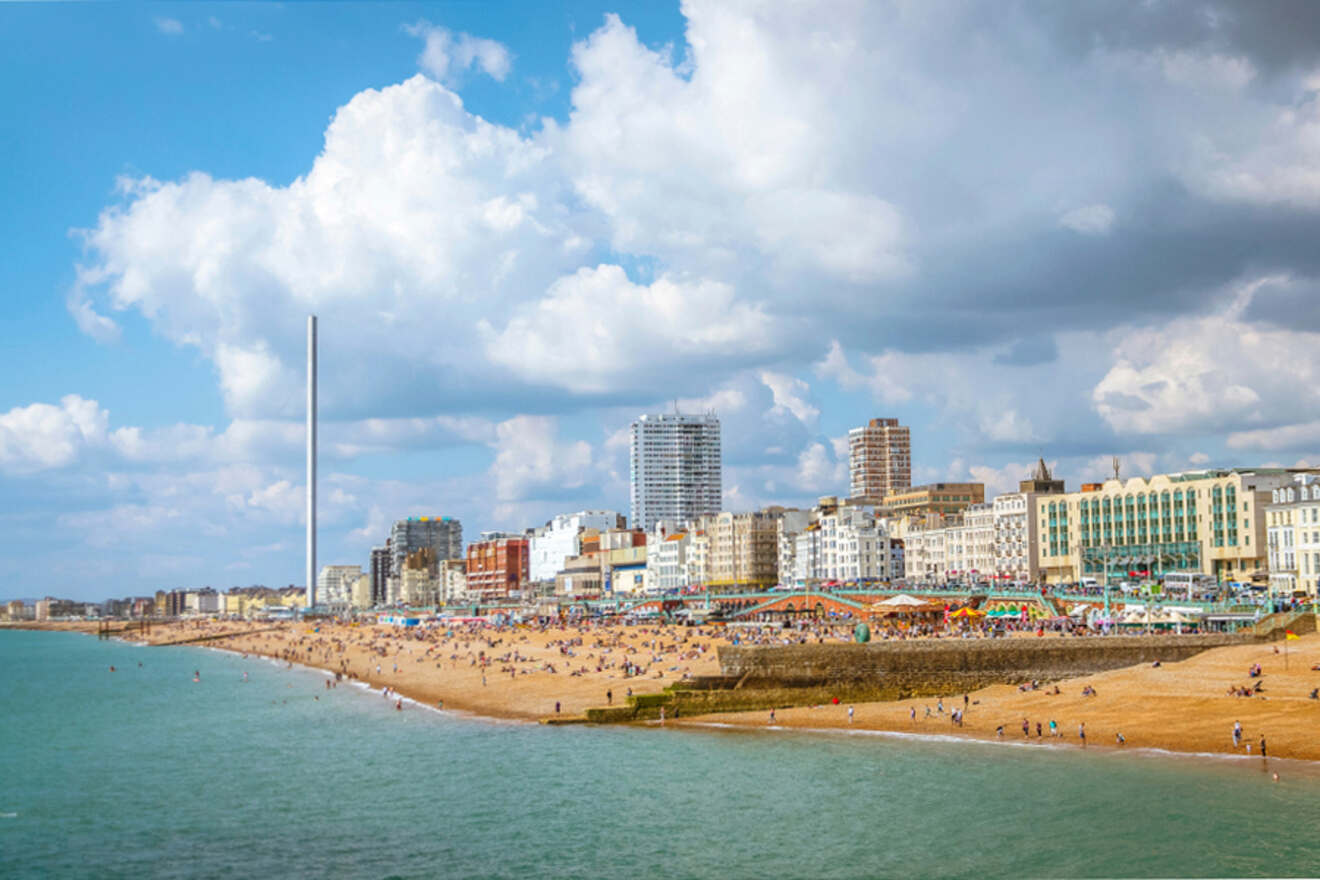 Beachfront cityscape with sandy shore, people sunbathing, and buildings in the background under a partly cloudy sky. Tower structure visible to the left.