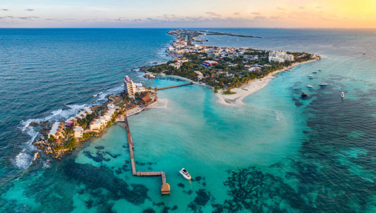 Aerial view of a narrow tropical island with buildings, surrounded by turquoise ocean waters, a lengthy dock, and distant horizon at sunset.