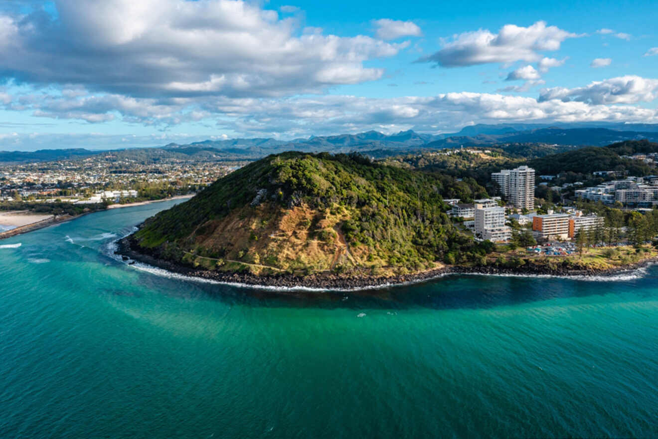 Aerial view of a coastal hill surrounded by ocean, with nearby buildings and distant mountains under a partly cloudy sky.