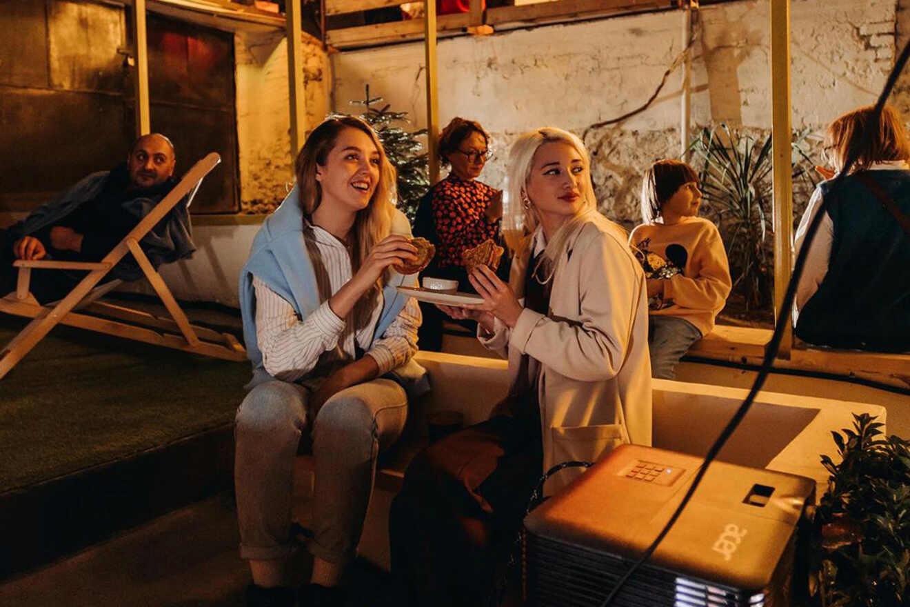 Two women sitting on a bench at a cozy indoor venue, holding plates of food. Several people are in the background. The scene is warmly lit, suggesting a relaxed, social atmosphere.