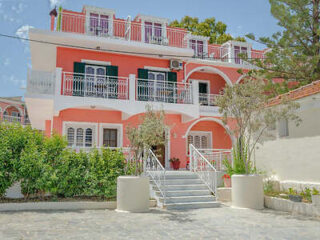 Three-story pink building with white trim, featuring balconies, arched windows, and a staircase leading to the entrance. Surrounded by greenery and trees.