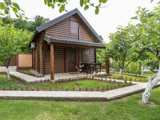 A small wooden cabin with a gabled roof, surrounded by grass and trees, with a stone pathway leading to the entrance.