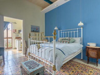 Bedroom with a white metal bed, ornate tiled flooring, blue accent wall, and vintage-style furniture. French doors lead to a balcony with a view.