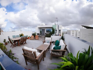 Rooftop patio with wooden furniture, green cushions, plants, and distant buildings under a cloudy sky.