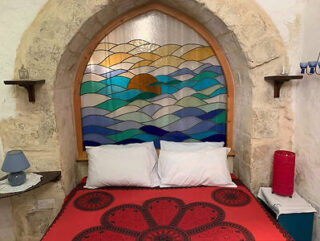 A bed with a red patterned cover is set against a stained glass backdrop depicting a sun setting over waves, framed by a stone arch.