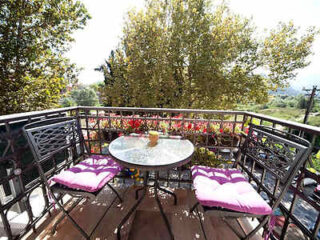 A small balcony with a round glass table, two metal chairs with pink cushions, overlooks green foliage and a distant landscape on a sunny day.