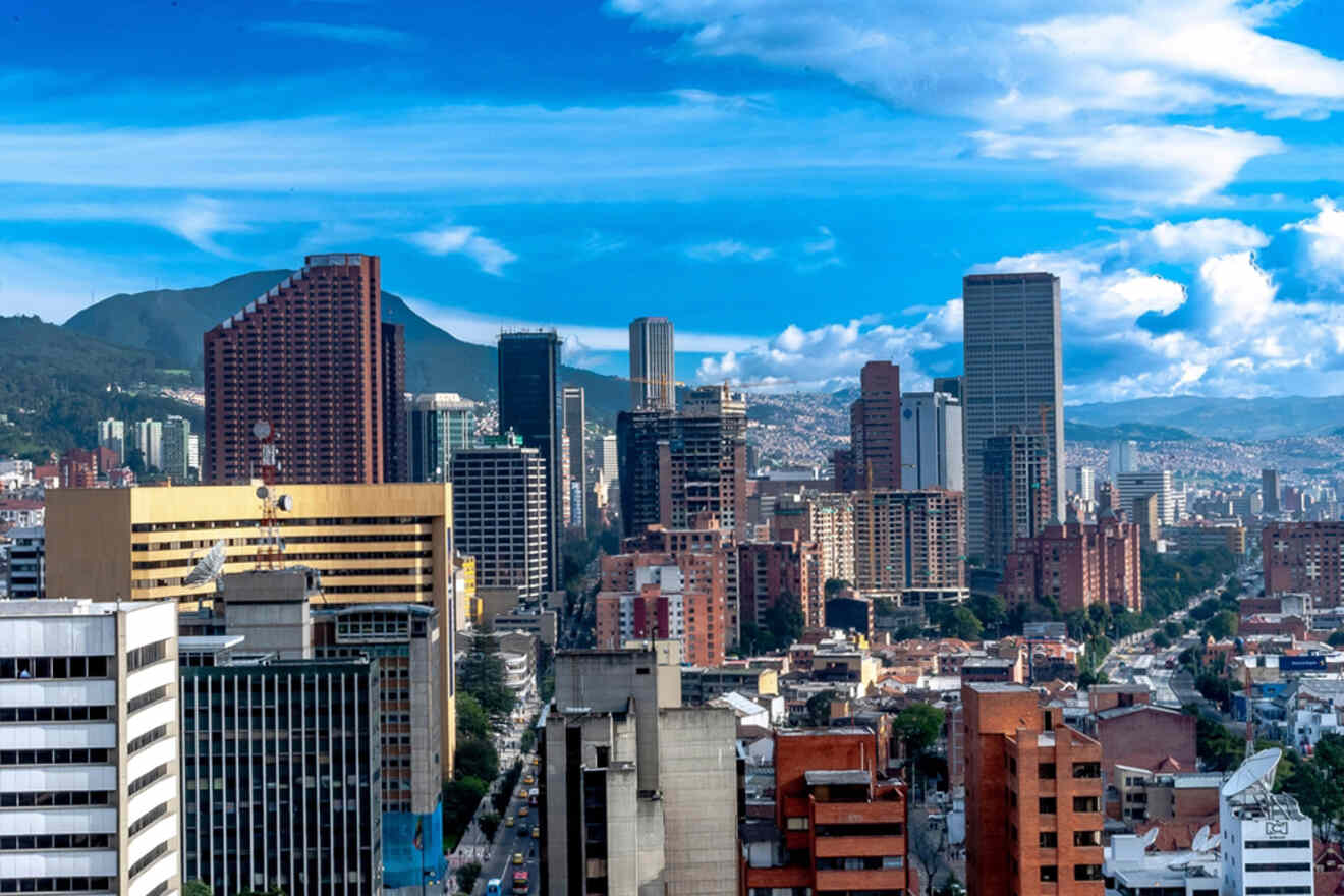 Skyline of a city with tall buildings under a blue sky, featuring mountains in the background.