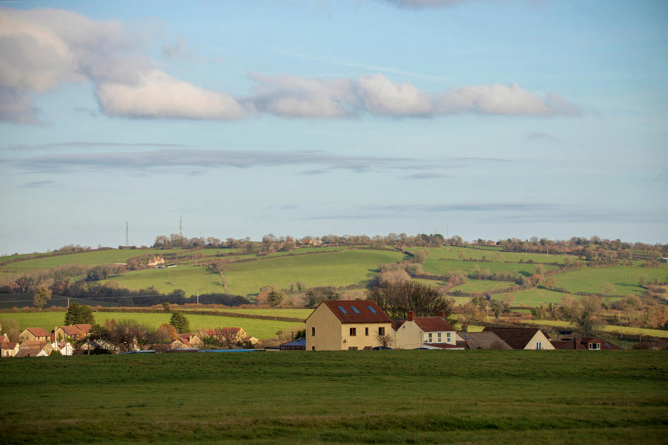 Countryside landscape featuring a small village with houses and cottages. Rolling green hills and fields stretch into the background under a partly cloudy sky.