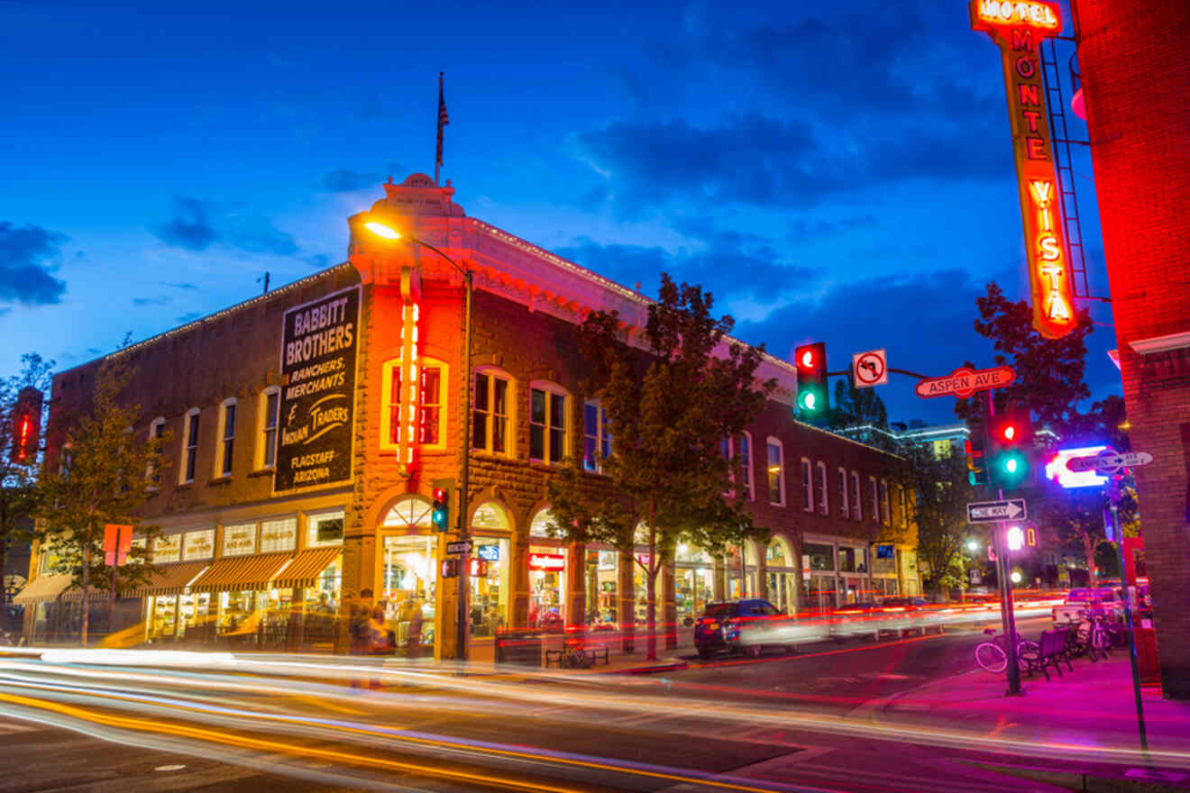 A bustling city intersection at dusk with light trails from passing cars. A historic building is illuminated, and bright neon signs are visible.