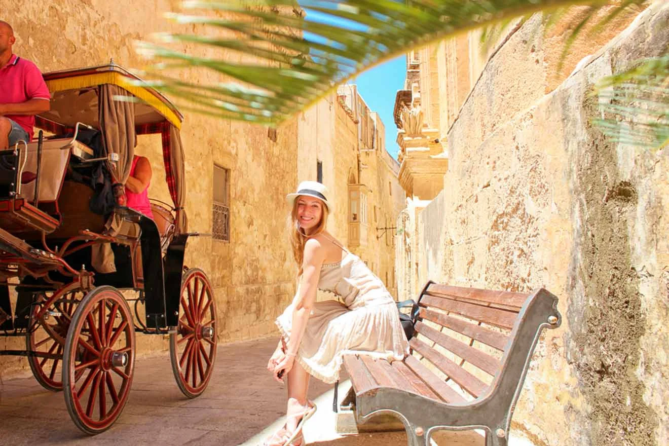 A woman in a white dress and hat sits on a bench in a sunlit, narrow street. A horse-drawn carriage with a driver is in the background.