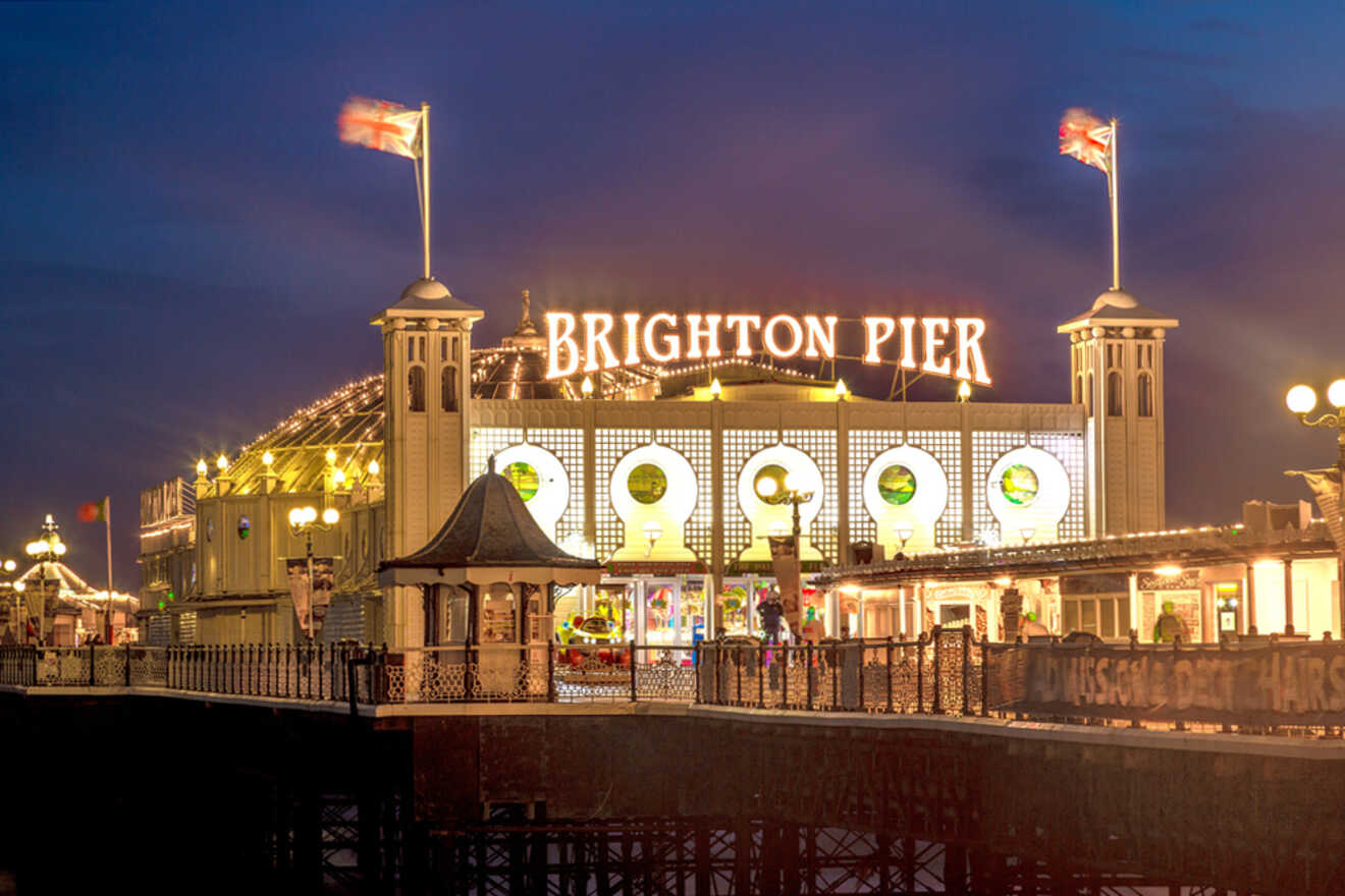 Brighton Pier at night, illuminated with lights and flanked by British flags, against a dark sky.