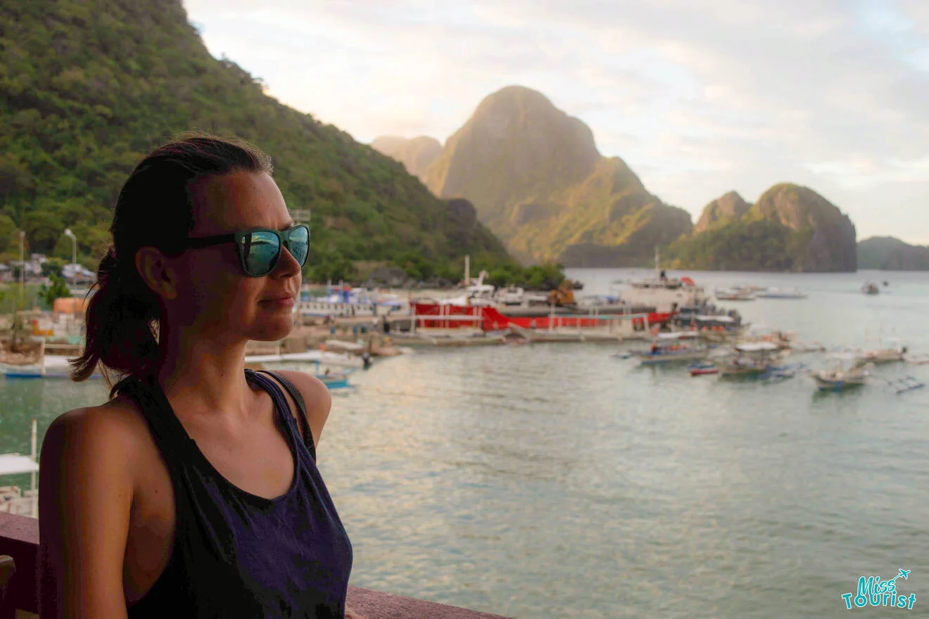 The writer of the post wearing sunglasses looks at a coastal landscape with boats and hills at sunset.