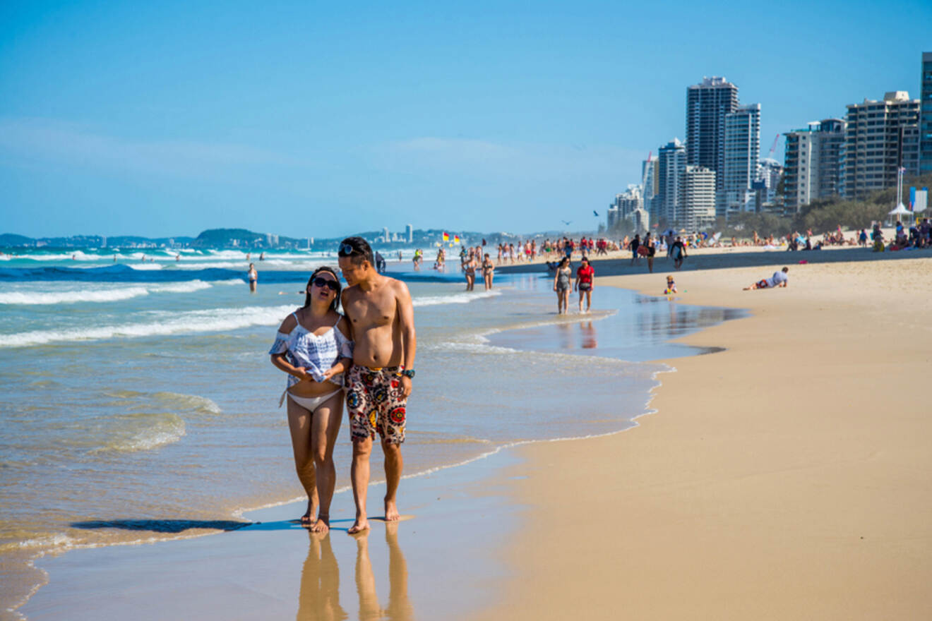 A couple walks on a sandy beach by the ocean, with city skyscrapers in the background on a sunny day.