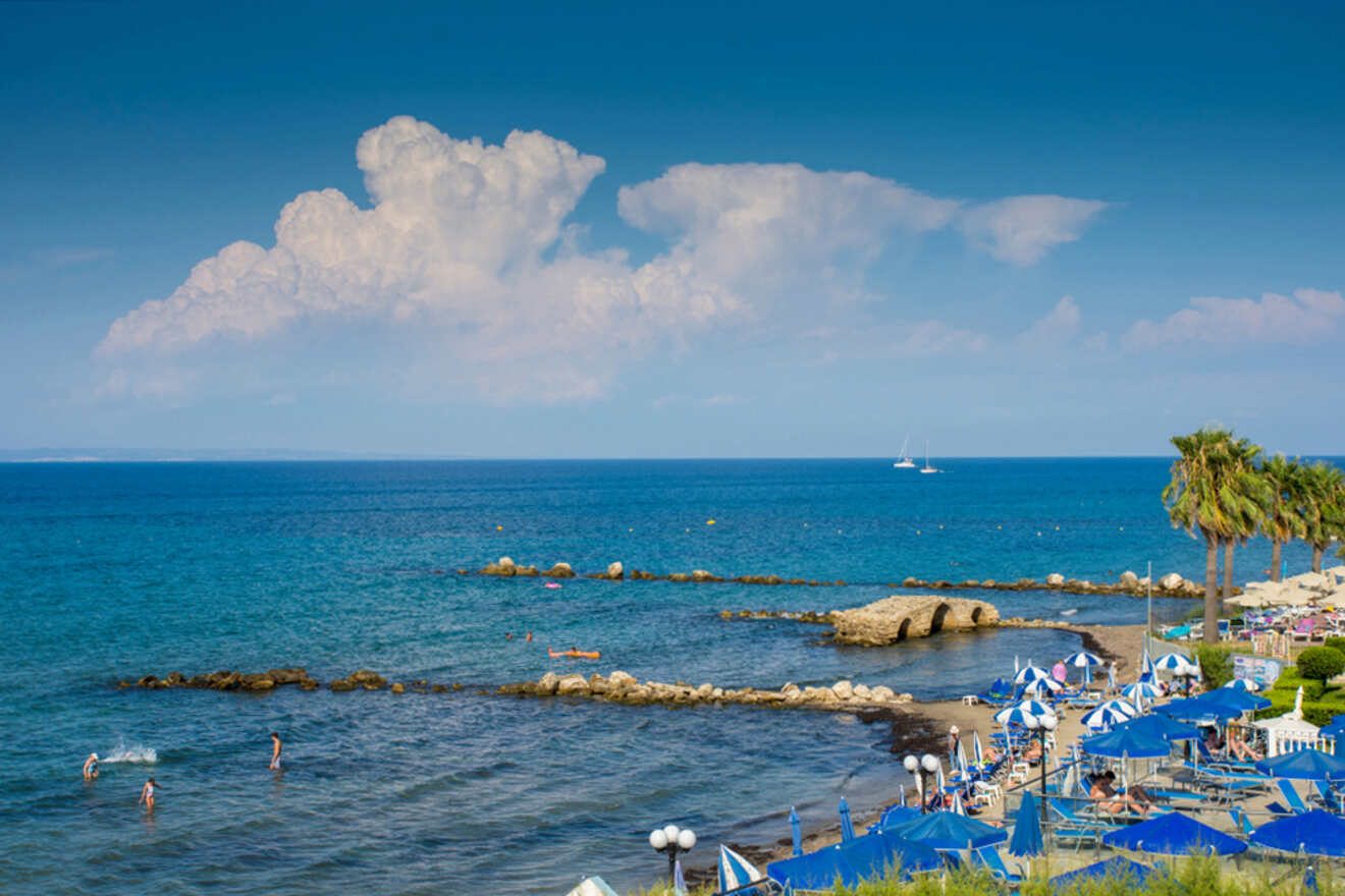 Beach scene with people swimming and lounging under blue-and-white umbrellas, a rocky shoreline, palm trees, and blue sea with a boat in the distance under a partly cloudy sky.