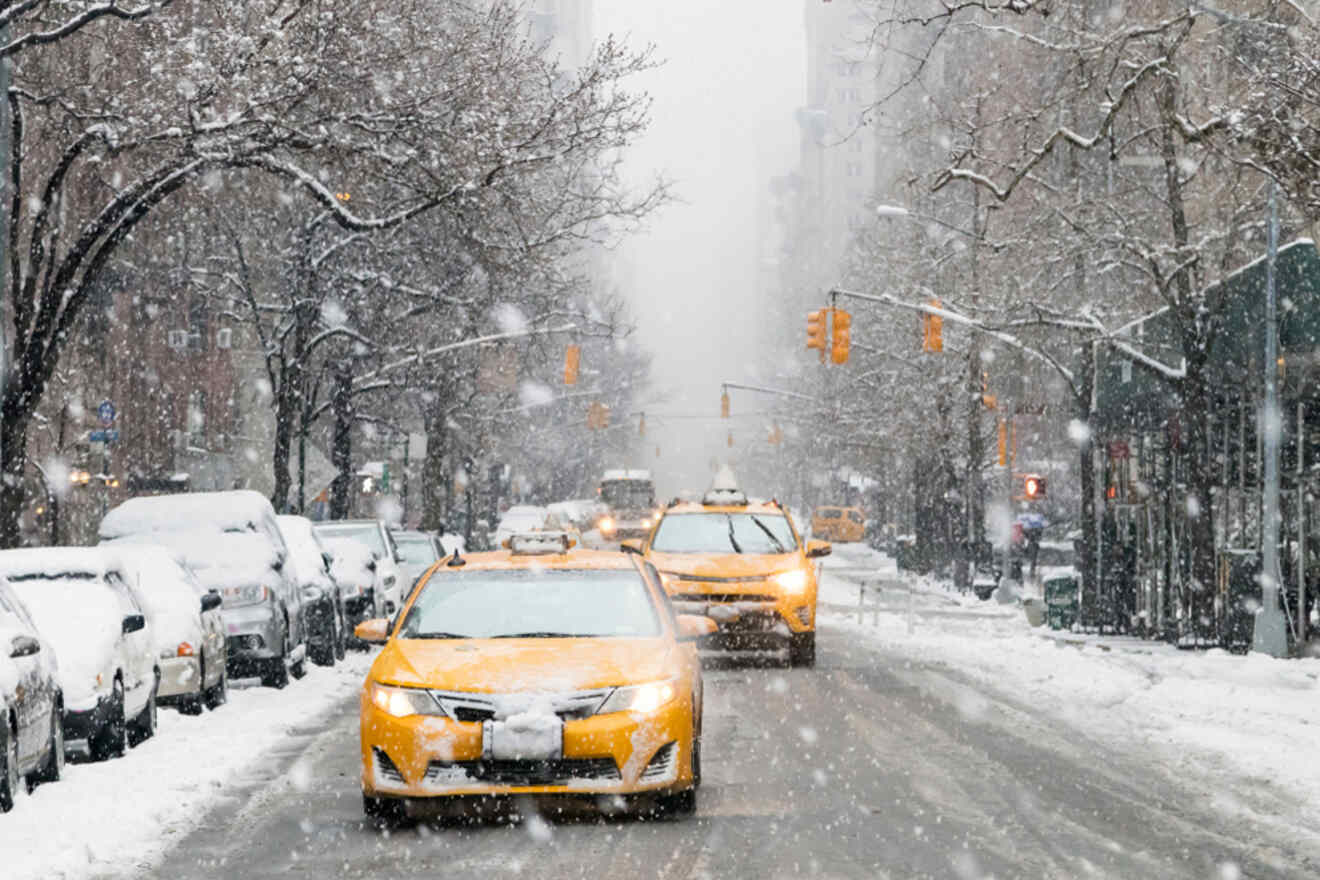 Snow-covered street with two yellow taxis driving, surrounded by parked cars and snow-dusted trees. Snowfall obscures distant buildings and traffic lights.