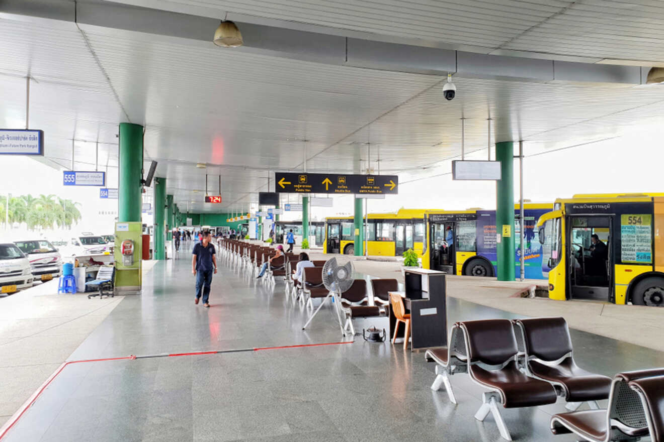 People walking in an airport terminal with seating areas, signs overhead, and parked buses in the background.