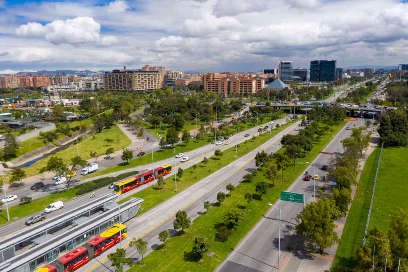 Aerial view of a cityscape with a highway, red buses, trees, and office buildings under a partly cloudy sky.