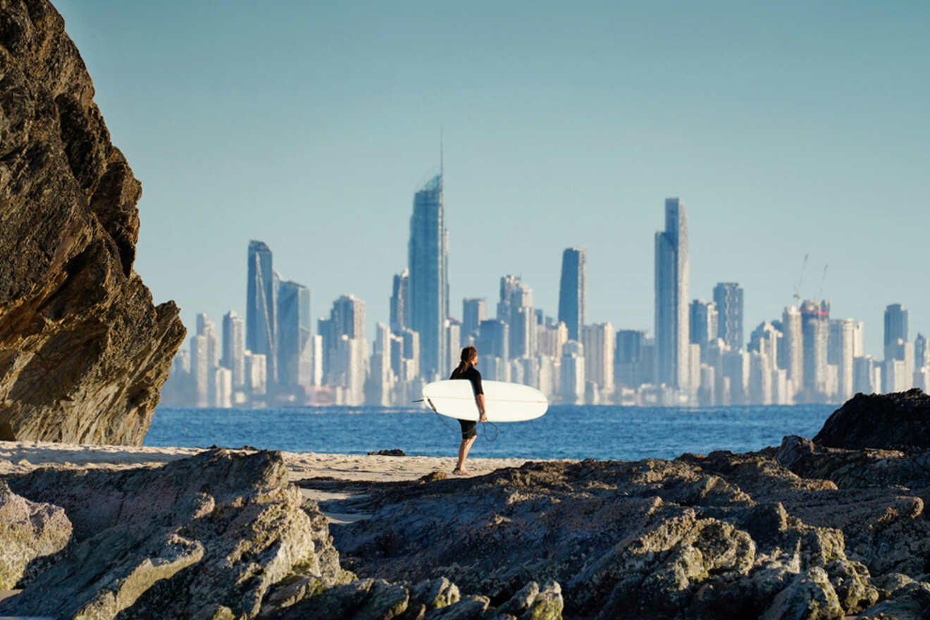 A person with a surfboard stands on a rocky shore, with a city skyline and tall buildings visible in the background.