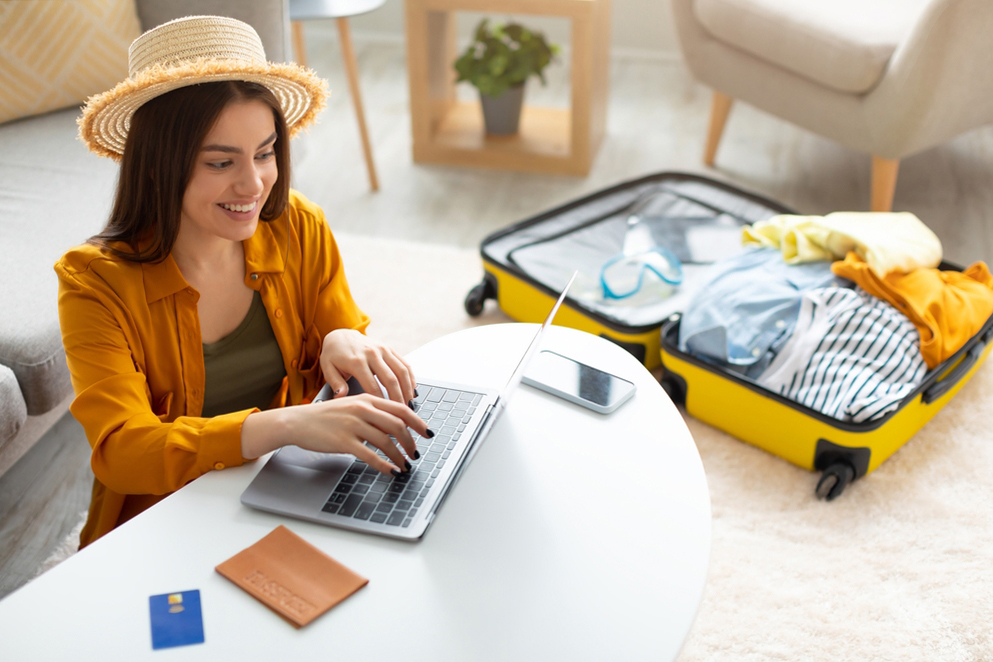 A woman with a straw hat uses a laptop on a white table. A credit card, phone, and passport are on the table. A yellow suitcase with clothes is open on the floor in the background.