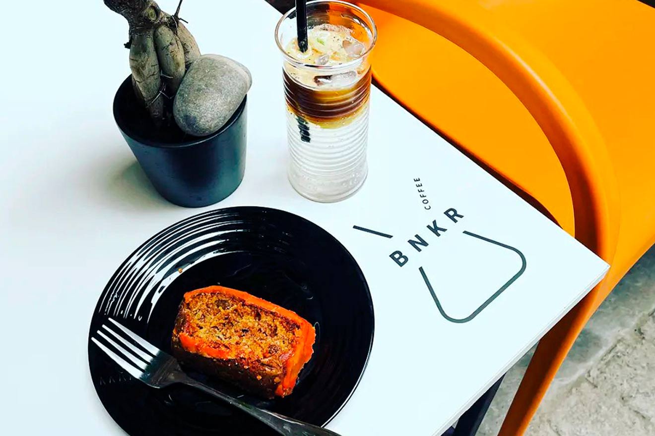 A slice of cake on a black plate, iced coffee in a tall glass, a small cactus in a pot, and a BNKR Coffee menu on an orange table.