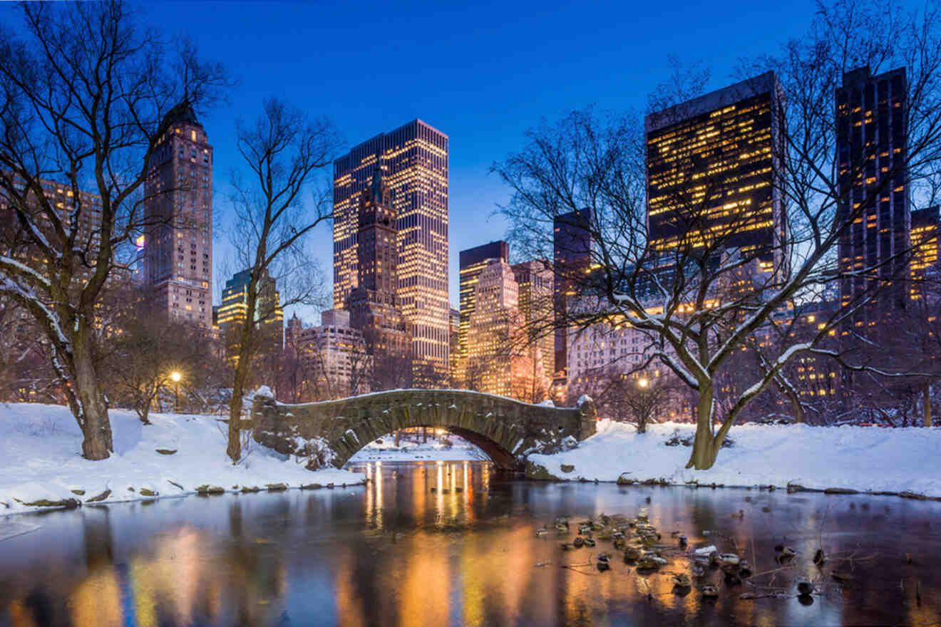 A snowy Central Park with a stone bridge and pond in the foreground, and illuminated skyscrapers in the background against a twilight sky.