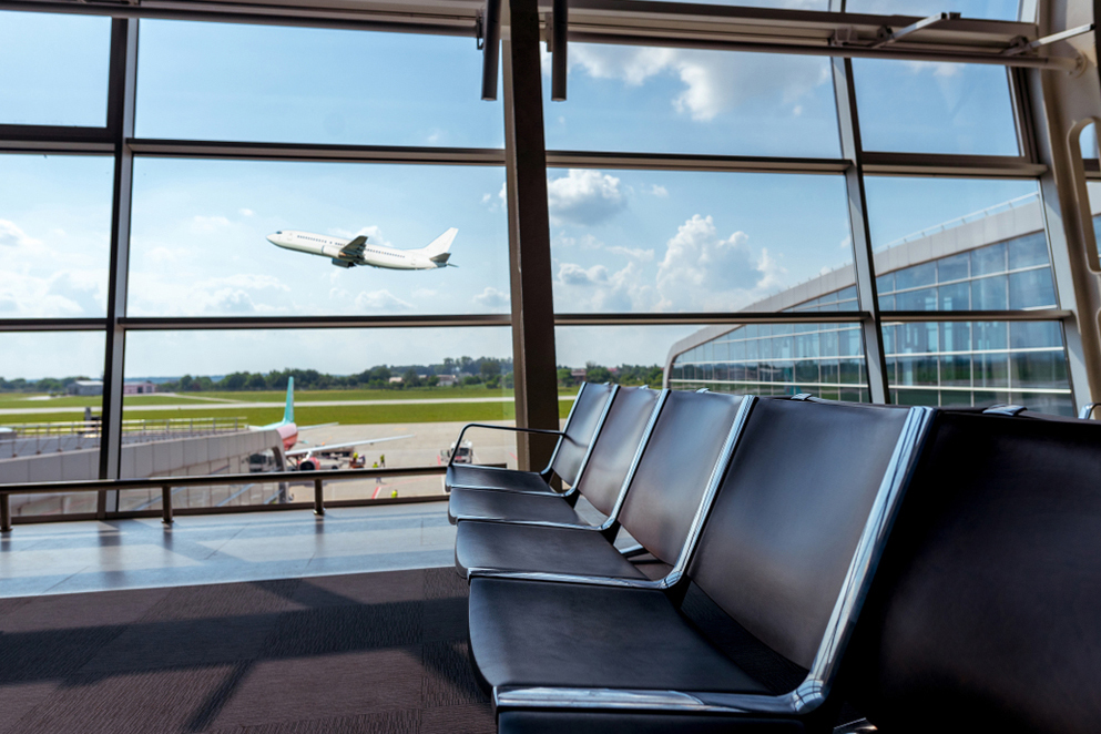 Empty airport seating area with a large window view of a plane taking off in the background under a blue sky with clouds.