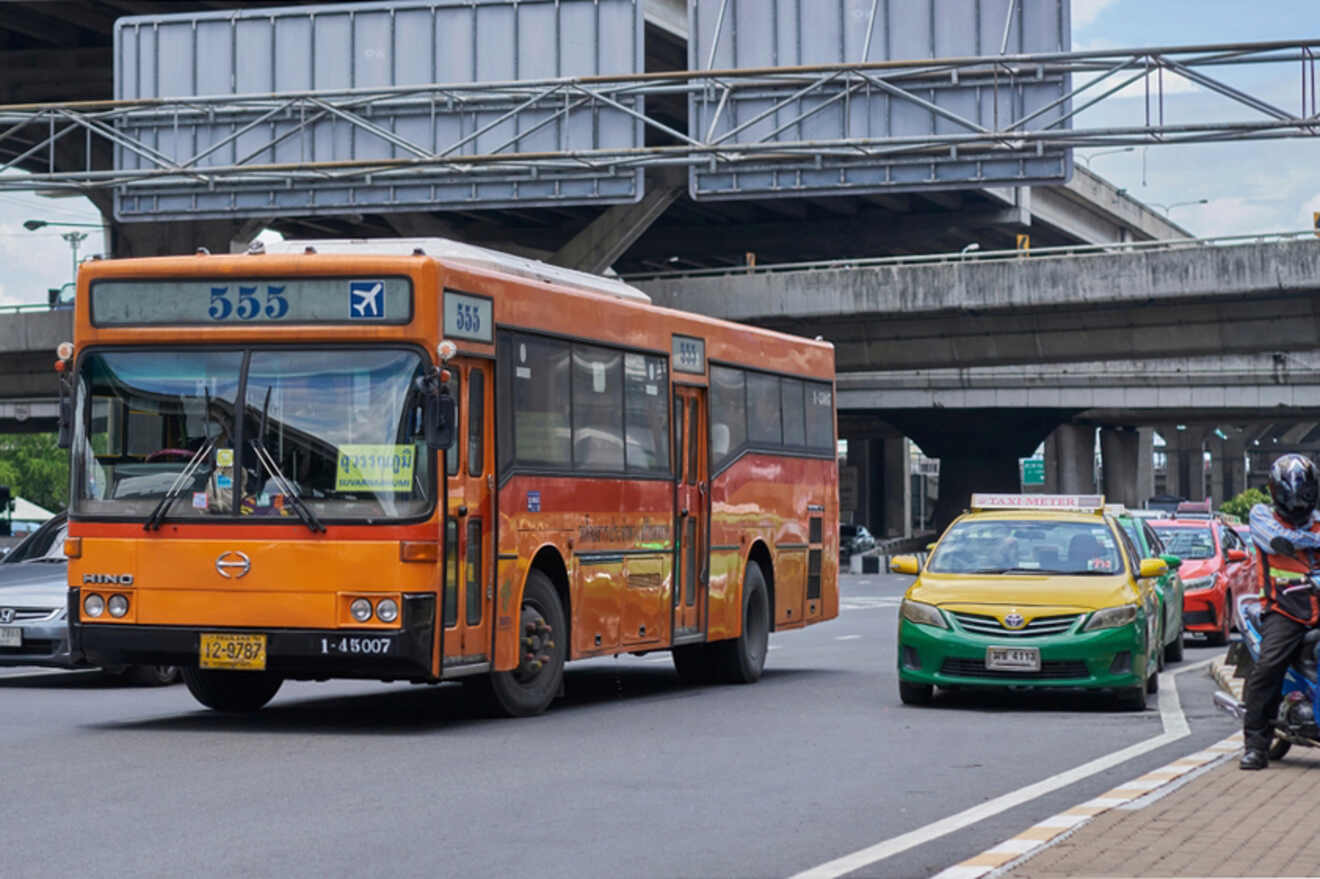 An orange city bus and a green taxi drive on a multi-lane road beneath an overpass. A motorbike is to the side.