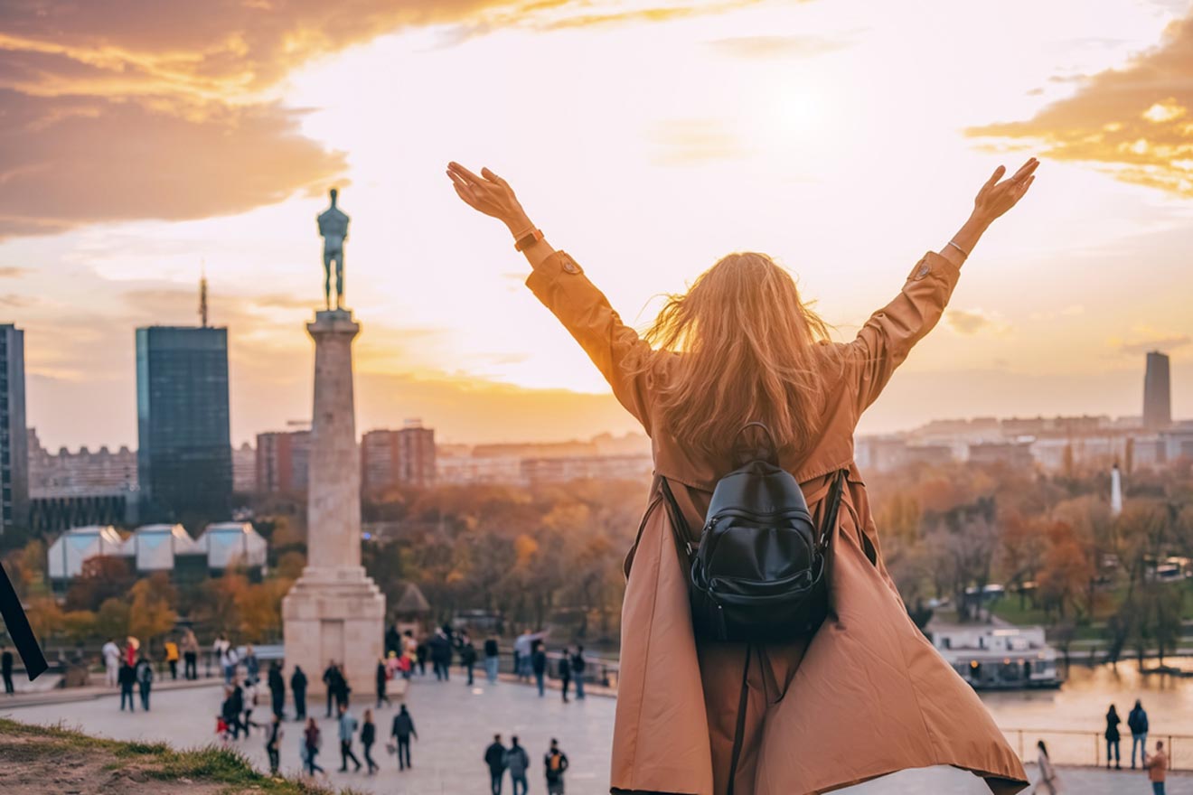 A person with outstretched arms stands on a high point, overlooking a cityscape at sunset with a statue and a crowd of people below.