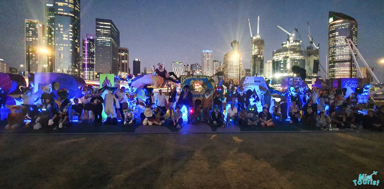 A large group of people in illuminated costumes gather outdoors at dusk. Skyscrapers are visible in the background.