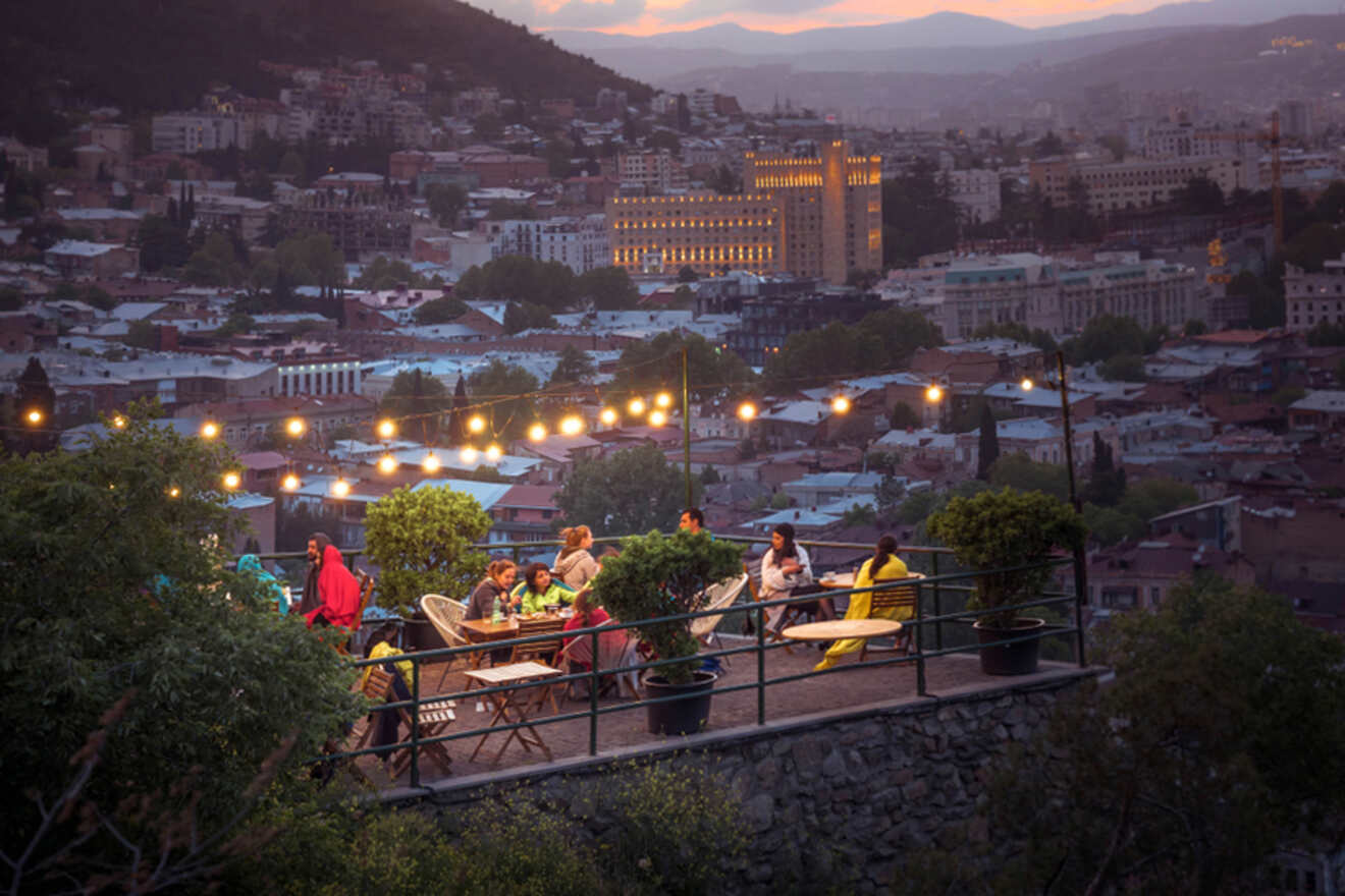 People sitting at outdoor restaurant tables under string lights, with a cityscape and hills in the background during twilight.