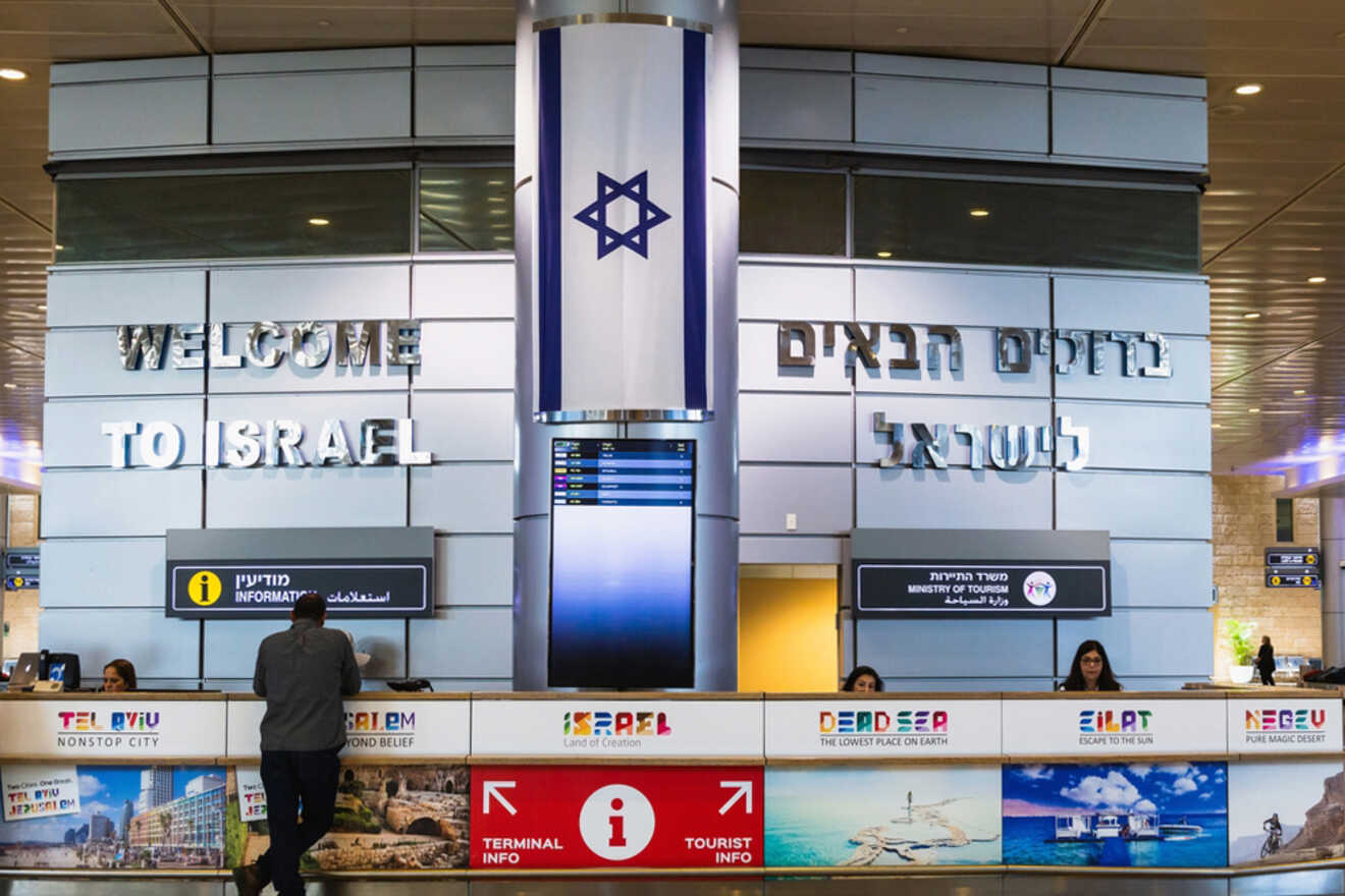 Interior of an airport terminal with "Welcome to Israel" on the wall, a flag, and a central information desk.