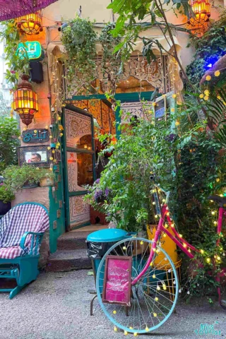 A colorful cafe entrance with ornate decorations, surrounded by lush greenery and hanging lanterns. A vintage bicycle with string lights is displayed in front.