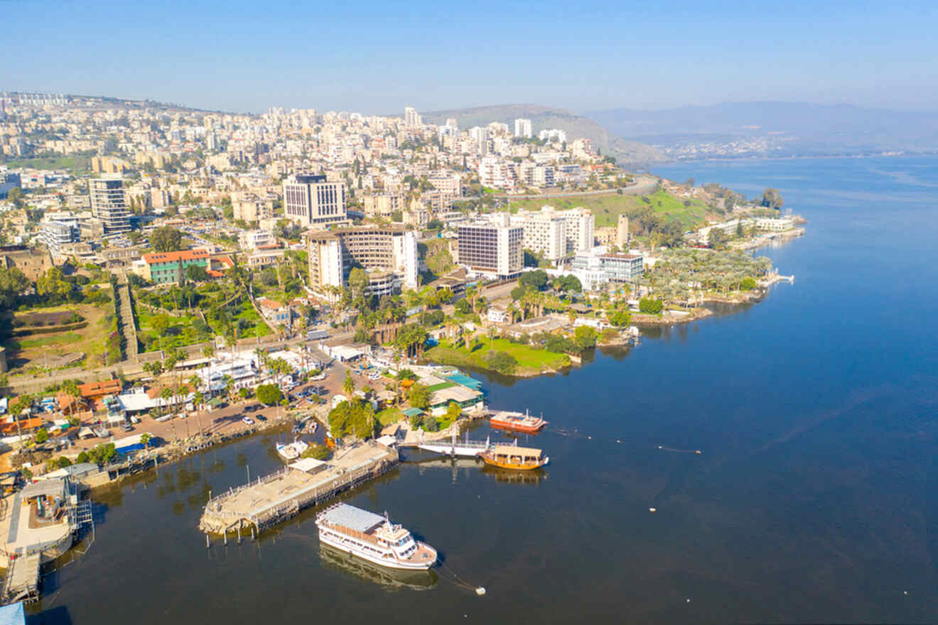 Aerial view of a coastal city with buildings and hills, featuring a harbor with several boats on a calm body of water.