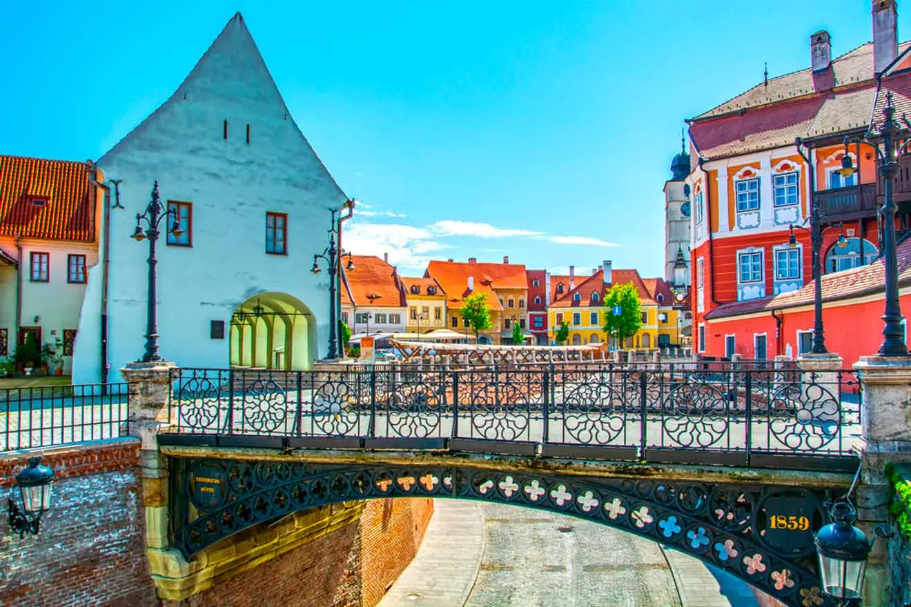 A picturesque European street featuring colorful historic buildings, a decorative iron bridge, and a clear blue sky.