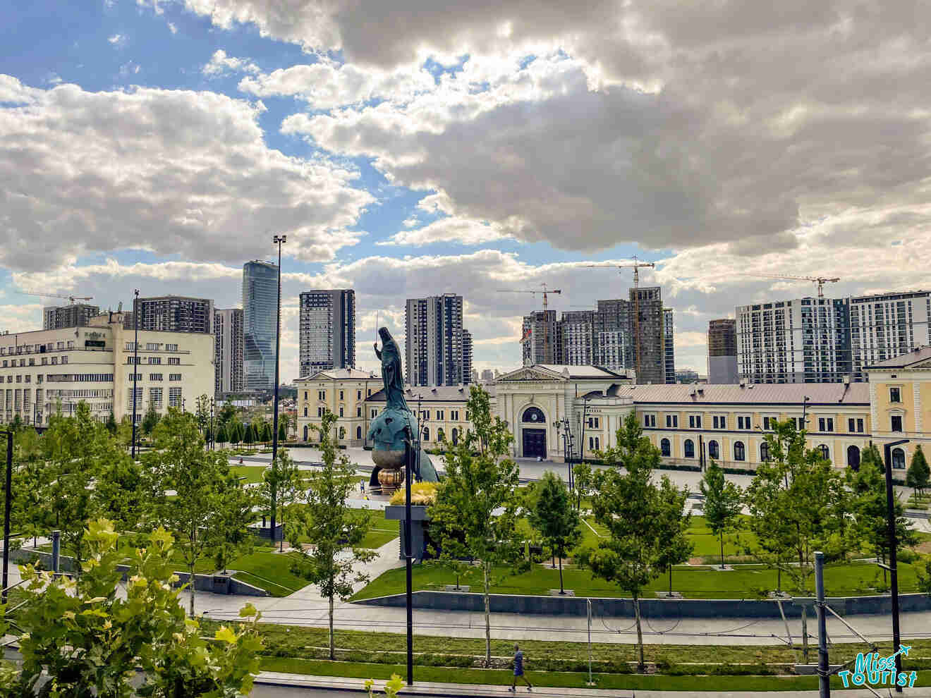 Cityscape with modern high-rise buildings behind a green park featuring a sculpture, under a partly cloudy sky.