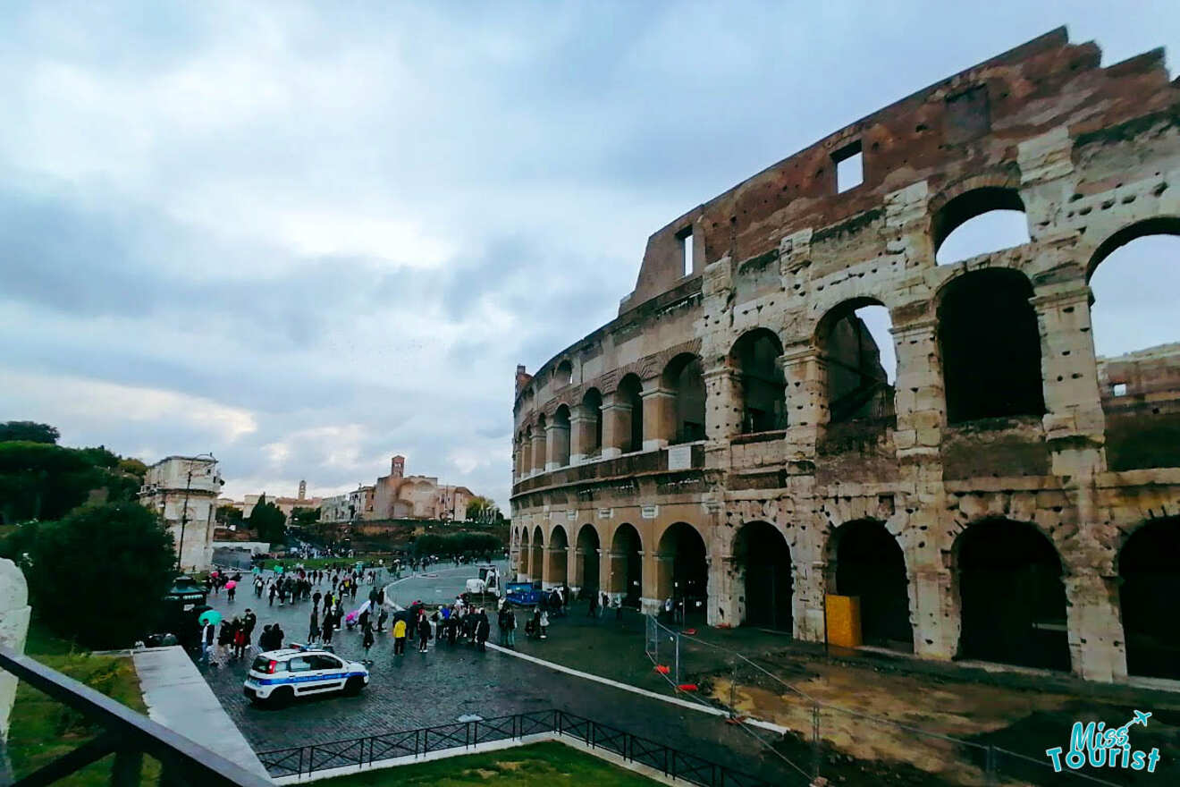 View of the Colosseum in Rome with people walking nearby and a police car parked on the street. Cloudy sky in the background.